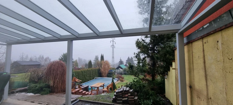 Pohled z hliníkové pergoly na zahradu s bazénem během deštivého dne
