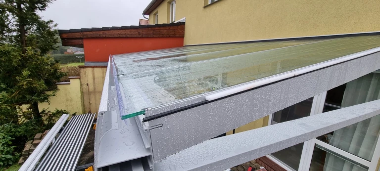 Detailní pohled na zastřešení hliníkové pergoly se skleněným výplňovým prvkem pokrytým kapkami deště.