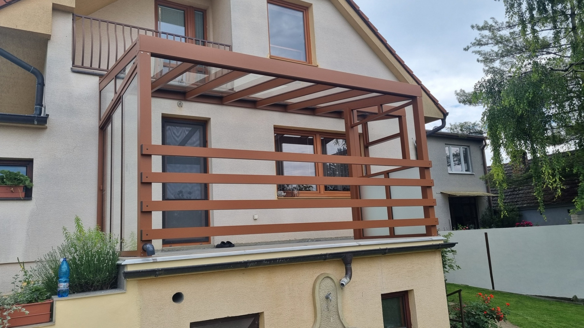 Hnědá atypická hliníková pergola se skleněnou střechou a bočním stíněním na balkóně rodinného domu