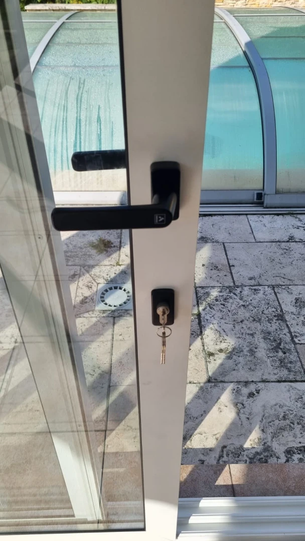 Detailní záběr na černou kliku a zámek s klíčem na bílých dveřích prosklené terasy.