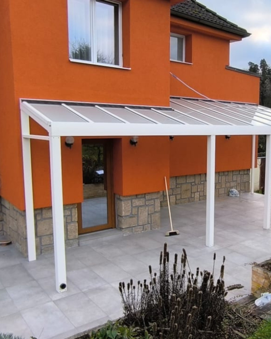 Moderní bílá hliníková pergola připevněná k oranžovému domu se skleněnou střechou a dlažbou