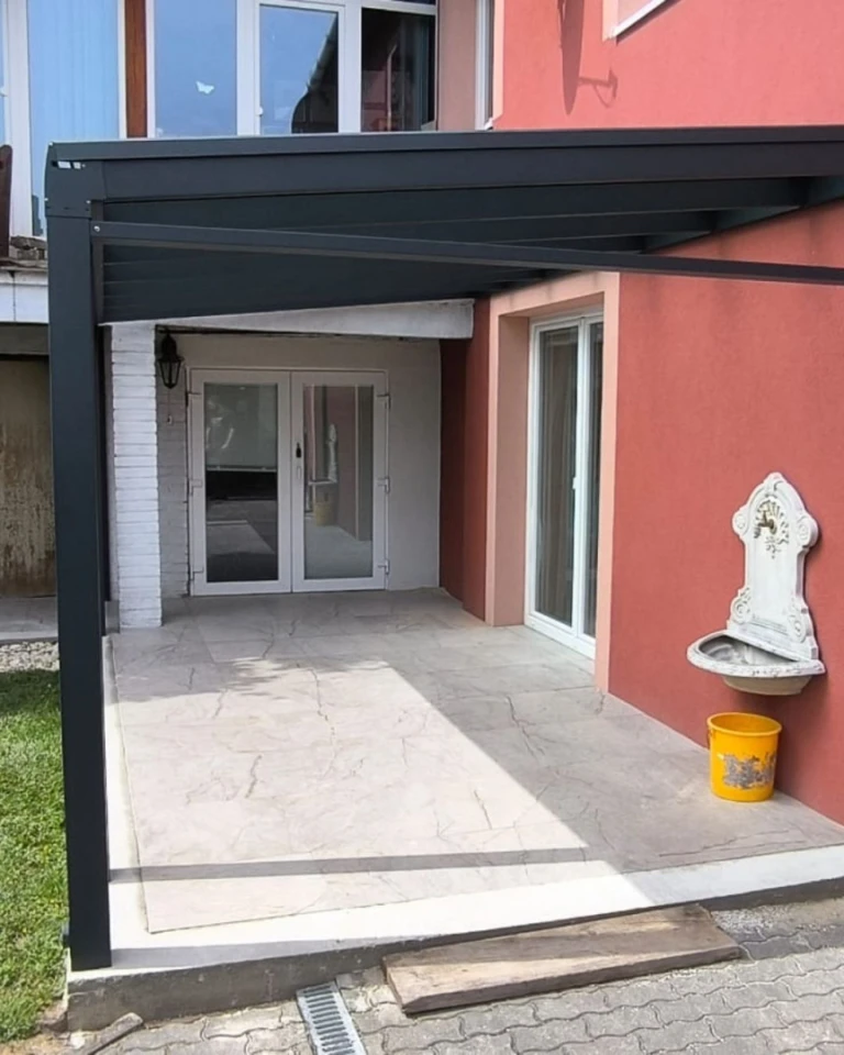 Moderní hliníková konstrukce pergoly Robust u domu s červenou fasádou a terasou.