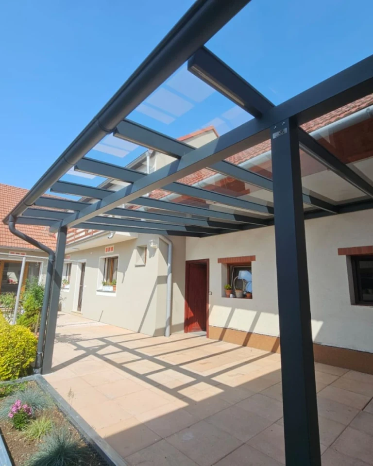 Moderní antracitová hliníková pergola se skleněnou střechou, připojená k rodinnému domu s dlážděnou terasou.