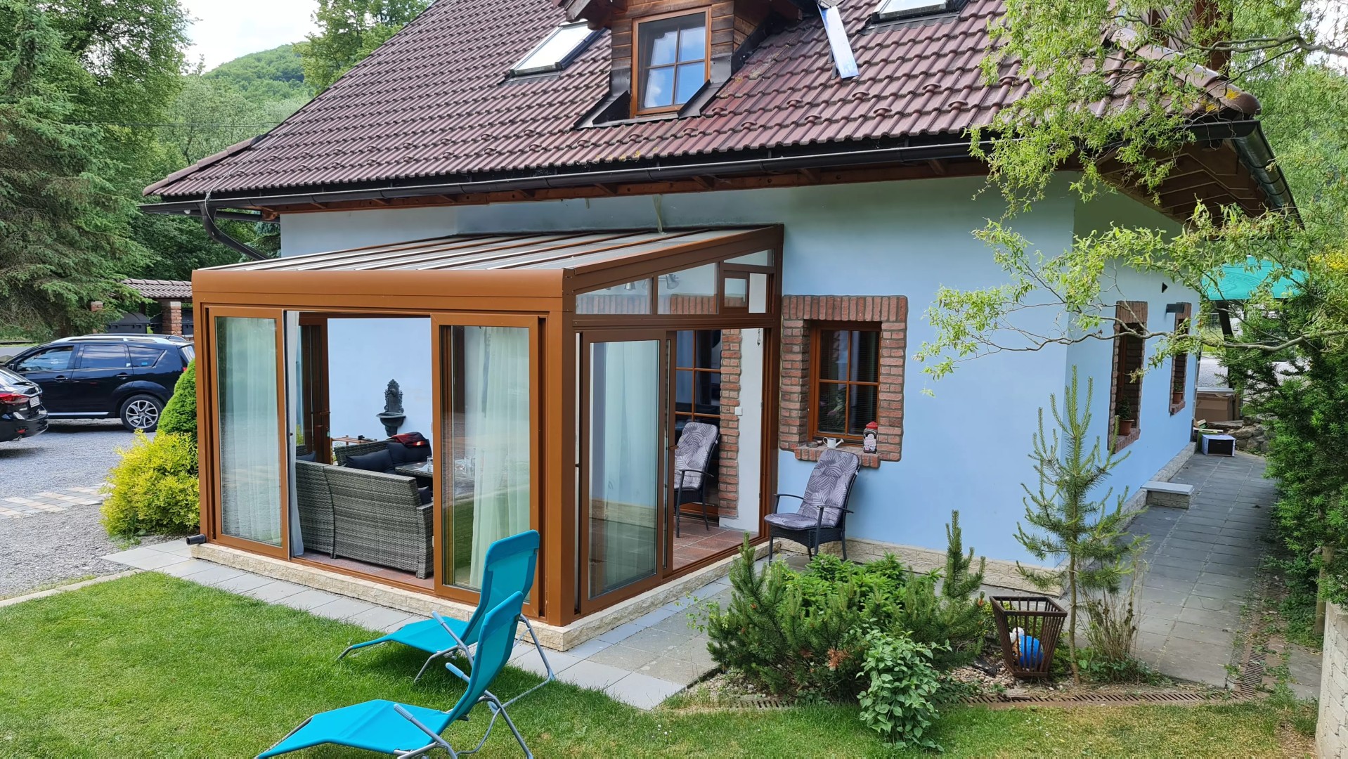 Prosklená hliníková pergola ROBUST v hnědé barvě, přistavěná k rodinnému domu, se zahradním nábytkem.