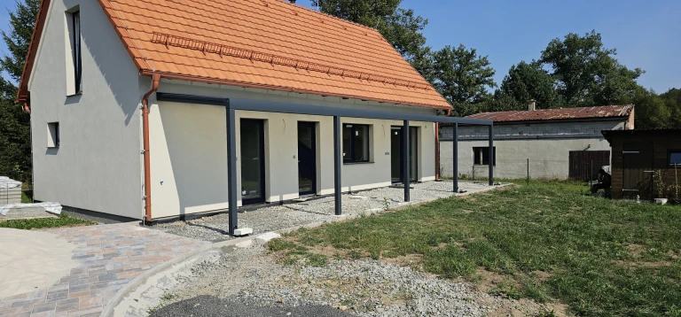 Moderní tmavě šedá hliníková konstrukce pergoly přistavěná k novostavbě rodinného domu s oranžovou střechou.