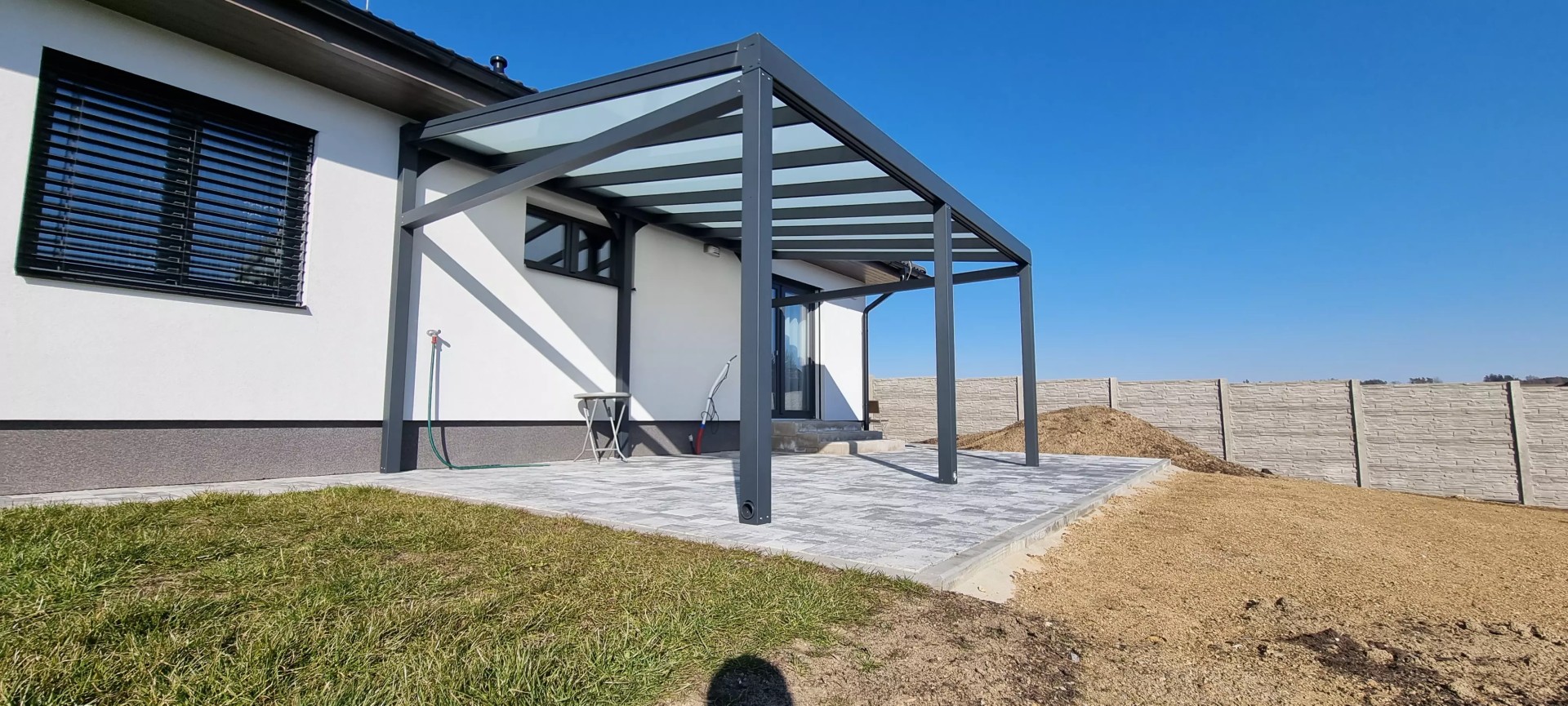 Moderní antracitová hliníková pergola se skleněnou střechou, přistavěná k rodinnému domu na dlážděné terase.