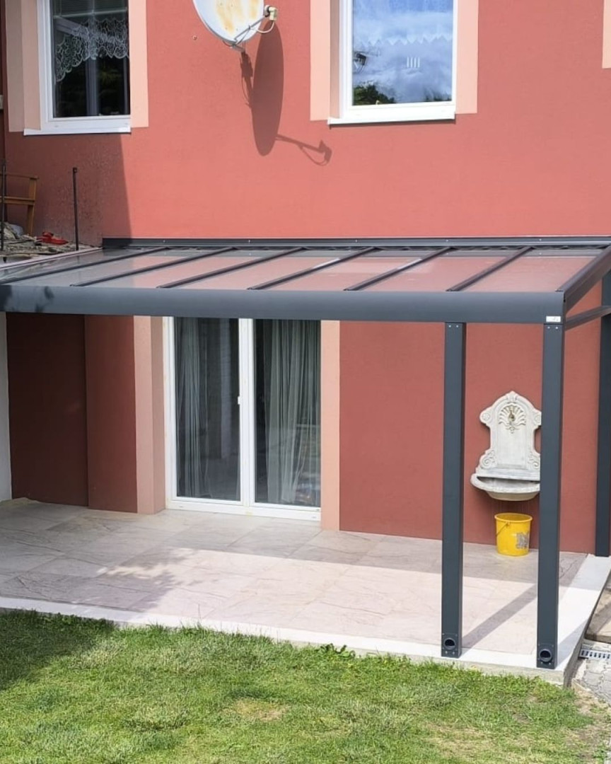 Moderní hliníková pergola se zasklenou střechou, připevněná ke zdi červeného domu nad terasou.