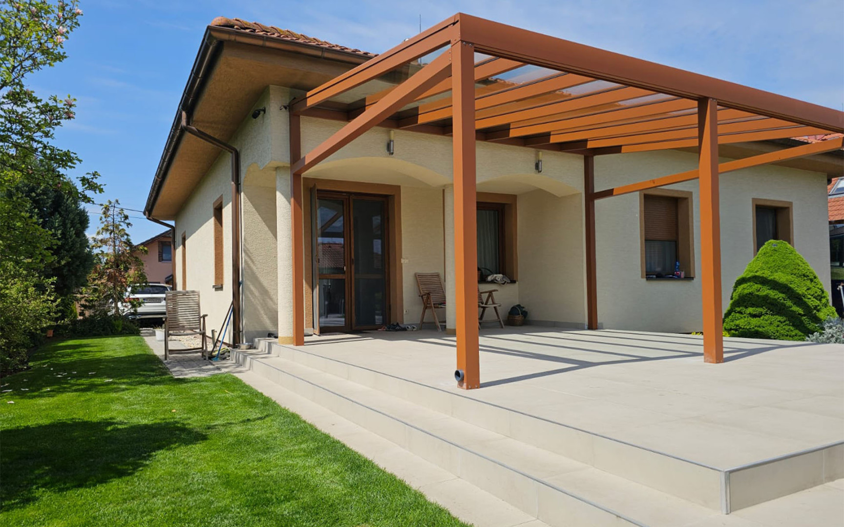 Moderní hliníková pergola v hnědé barvě se zasklenou střechou, připojená k rodinnému domu s prostornou terasou.