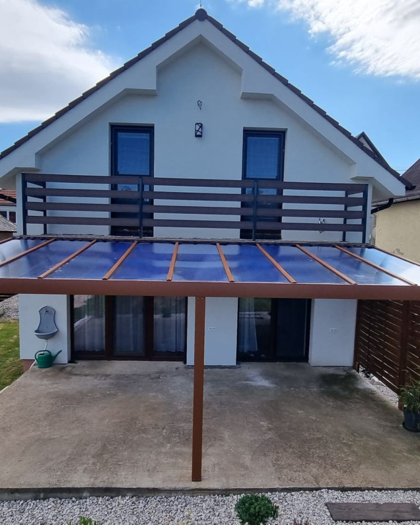 Hnědá hliníková pergola se skleněnou střechou přistavěná k rodinnému domu s balkonem.