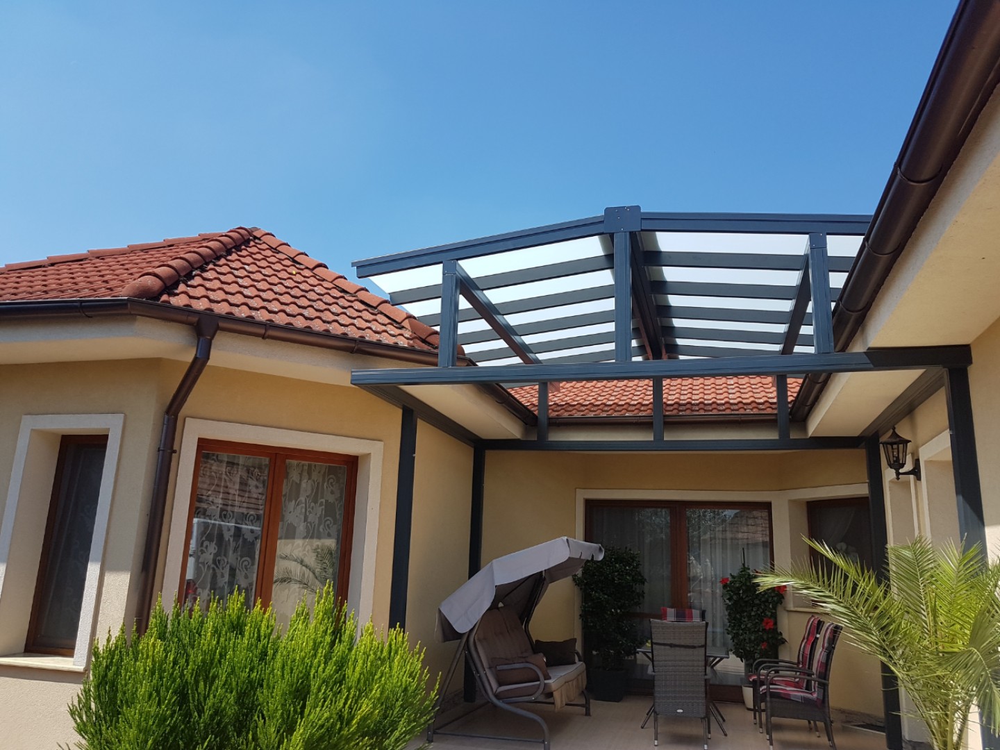 Moderní hliníková pergola ROBUST se zasklenou střechou, připojená k rodinnému domu, s terasovým nábytkem a houpačkou.