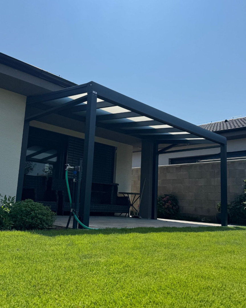Moderní tmavě šedá hliníková pergola připojená k domu se skleněnou střechou a upraveným trávníkem
