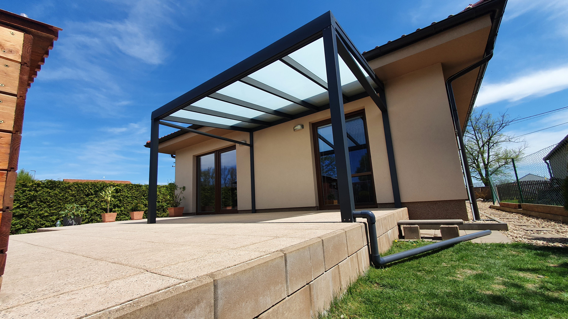 Moderní tmavě šedá hliníková pergola se skleněnou střechou, připevněná k rodinnému domu na dlážděné terase.