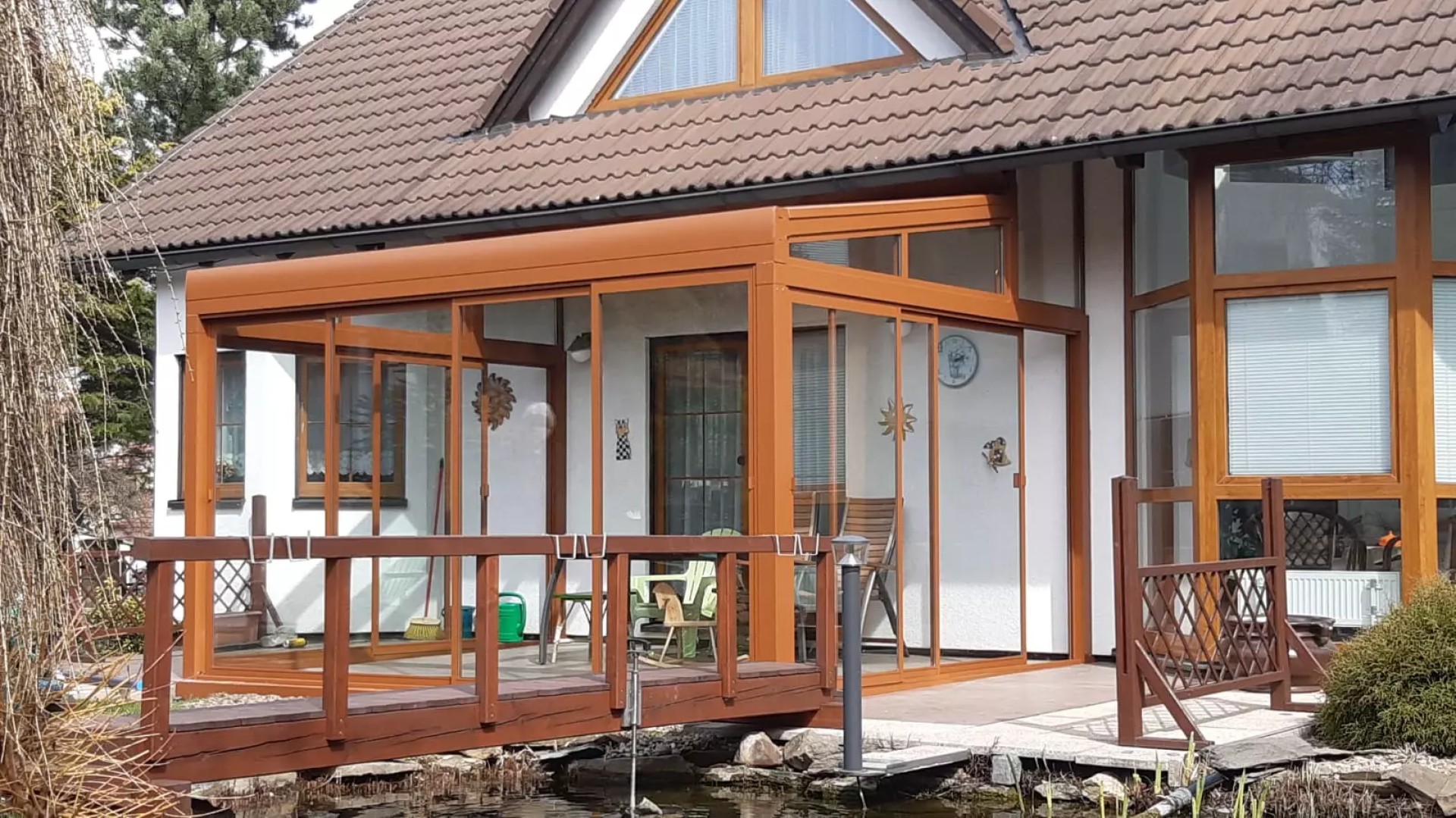 Prosklená hliníková pergola ROBUST v hnědé barvě u rodinného domu s dřevěným můstkem
