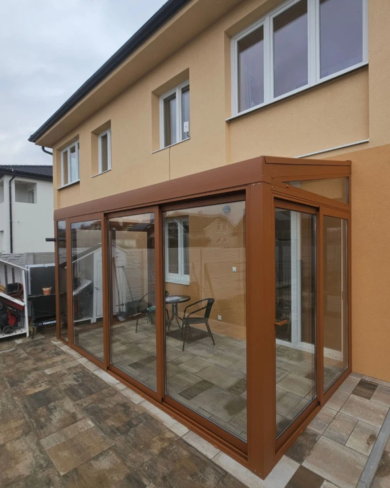 Moderní prosklená přístavba k rodinnému domu s hnědou konstrukcí a posuvnými dveřmi.