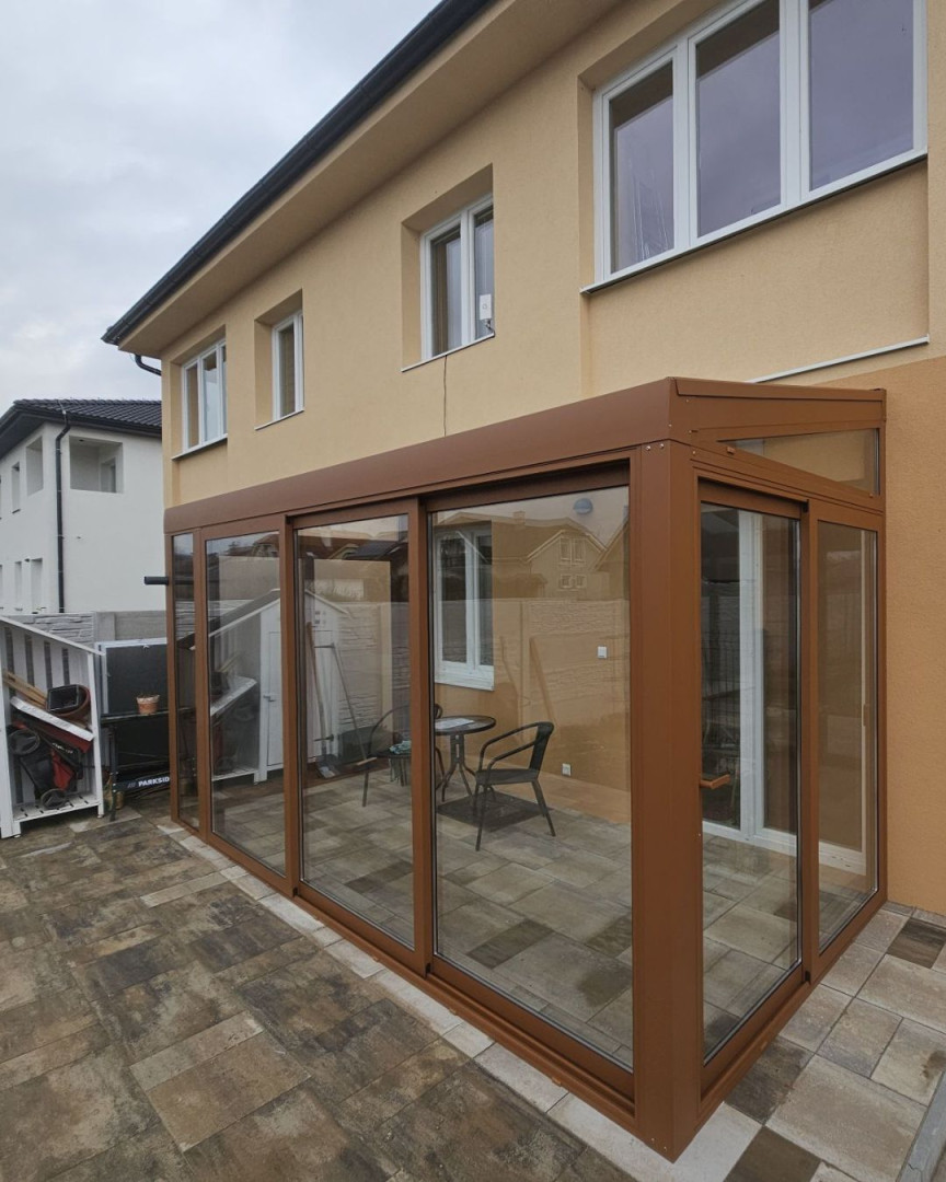 Moderní prosklená zimní zahrada s hnědou konstrukcí přistavěná k rodinnému domu