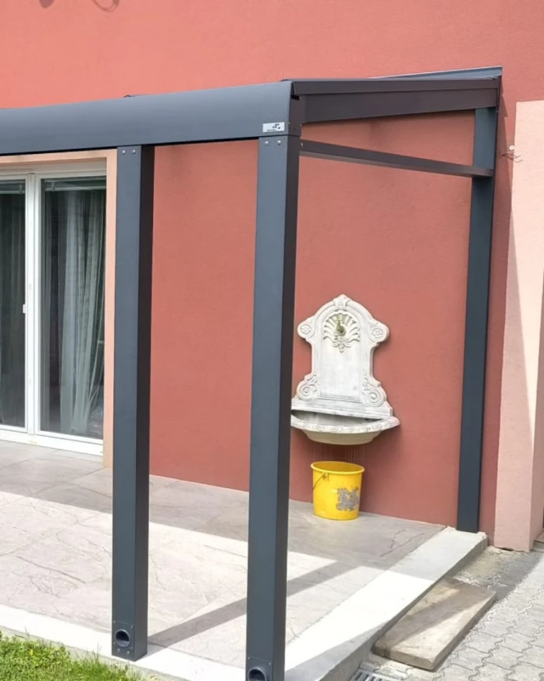 Detailní pohled na nosnou konstrukci a připojení hliníkové pergoly k fasádě domu s ozdobným umyvadlem.