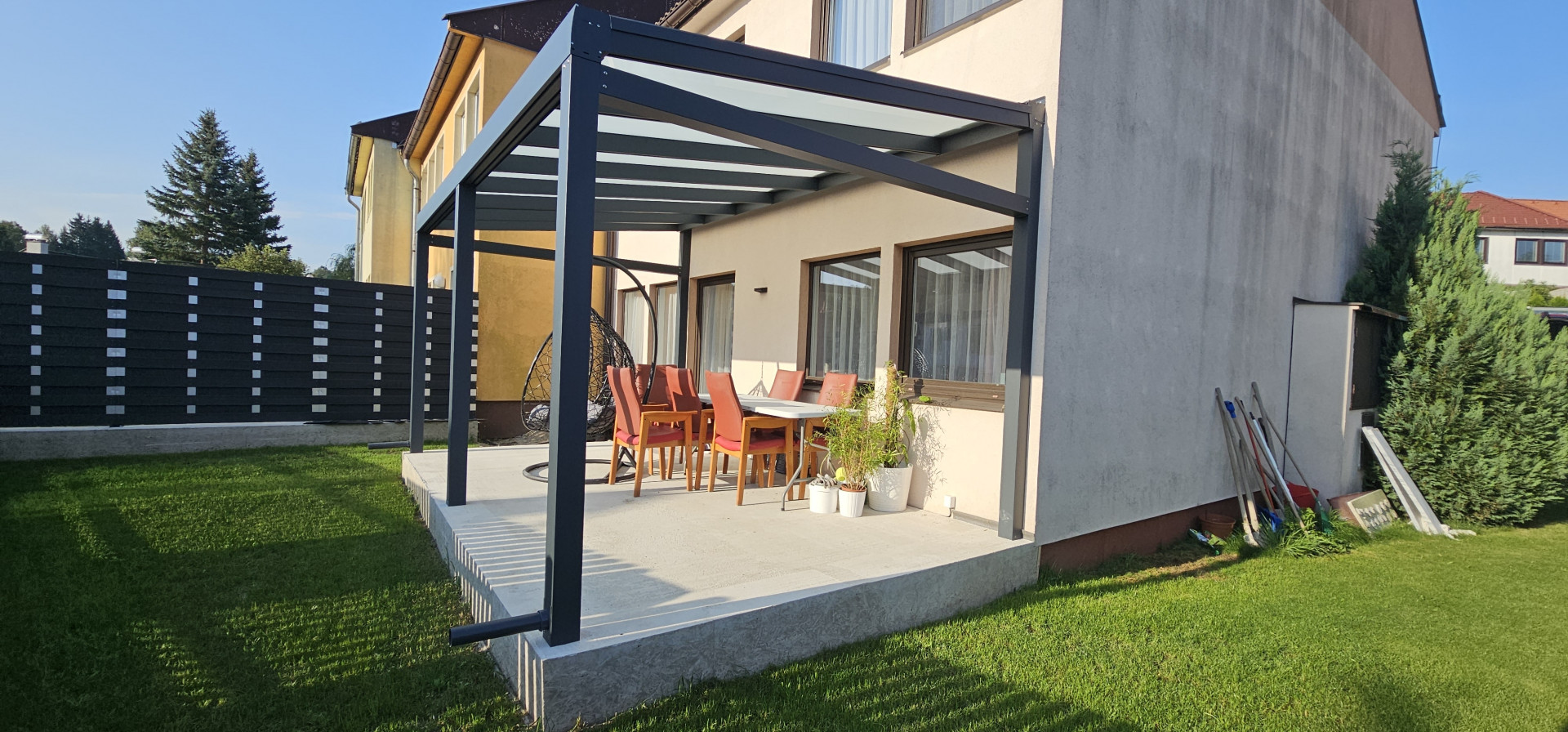 Moderní hliníková pergola se skleněnou střechou na betonové terase rodinného domu se zahradním nábytkem.