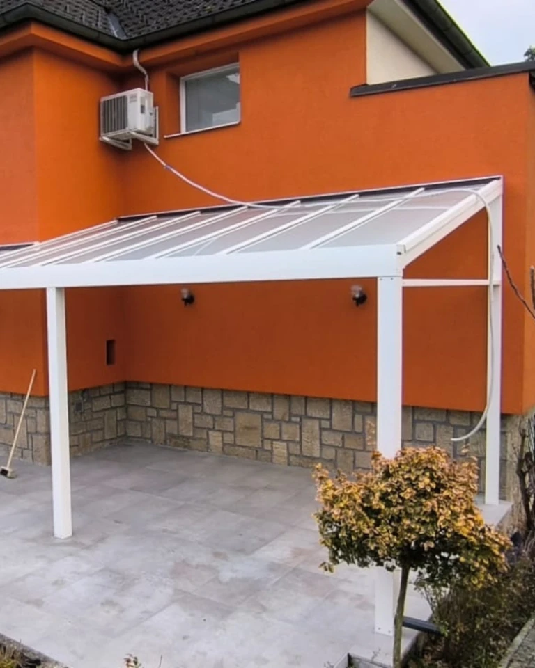 Bílá hliníková pergola se skleněnou střechou, připojená k oranžovému domu