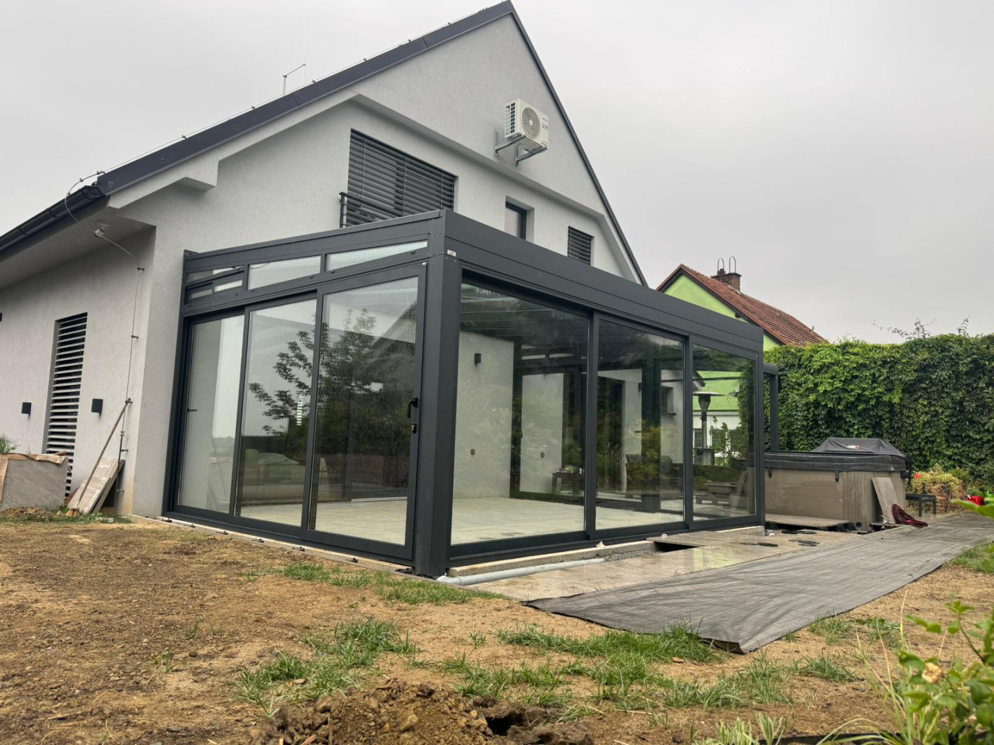 Moderní prosklená konstrukce připojená k rodinnému domu s tmavým rámem a posuvnými dveřmi.