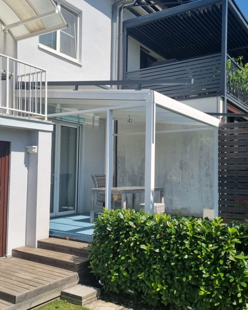 Bílá moderní hliníková pergola se skleněnou střechou u rodinného domu s terasovým nábytkem.