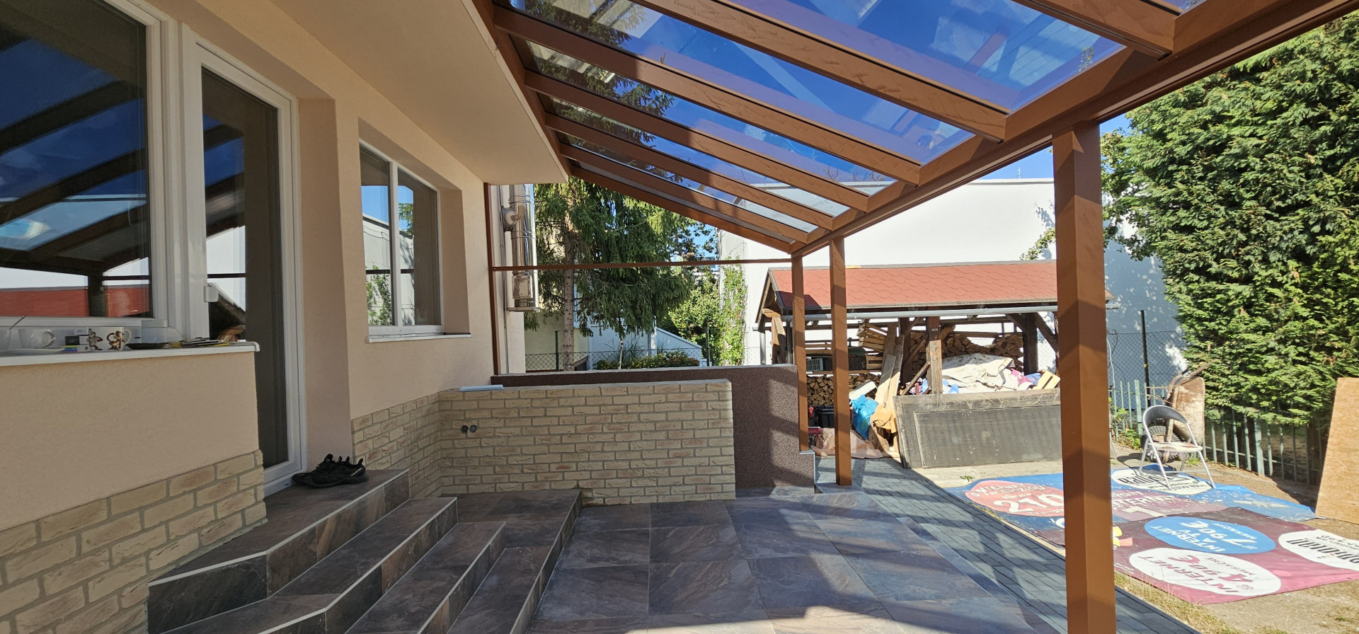 Hliníková pergola v dřevěném dekoru se skleněnou střechou, namontovaná na terase rodinného domu.