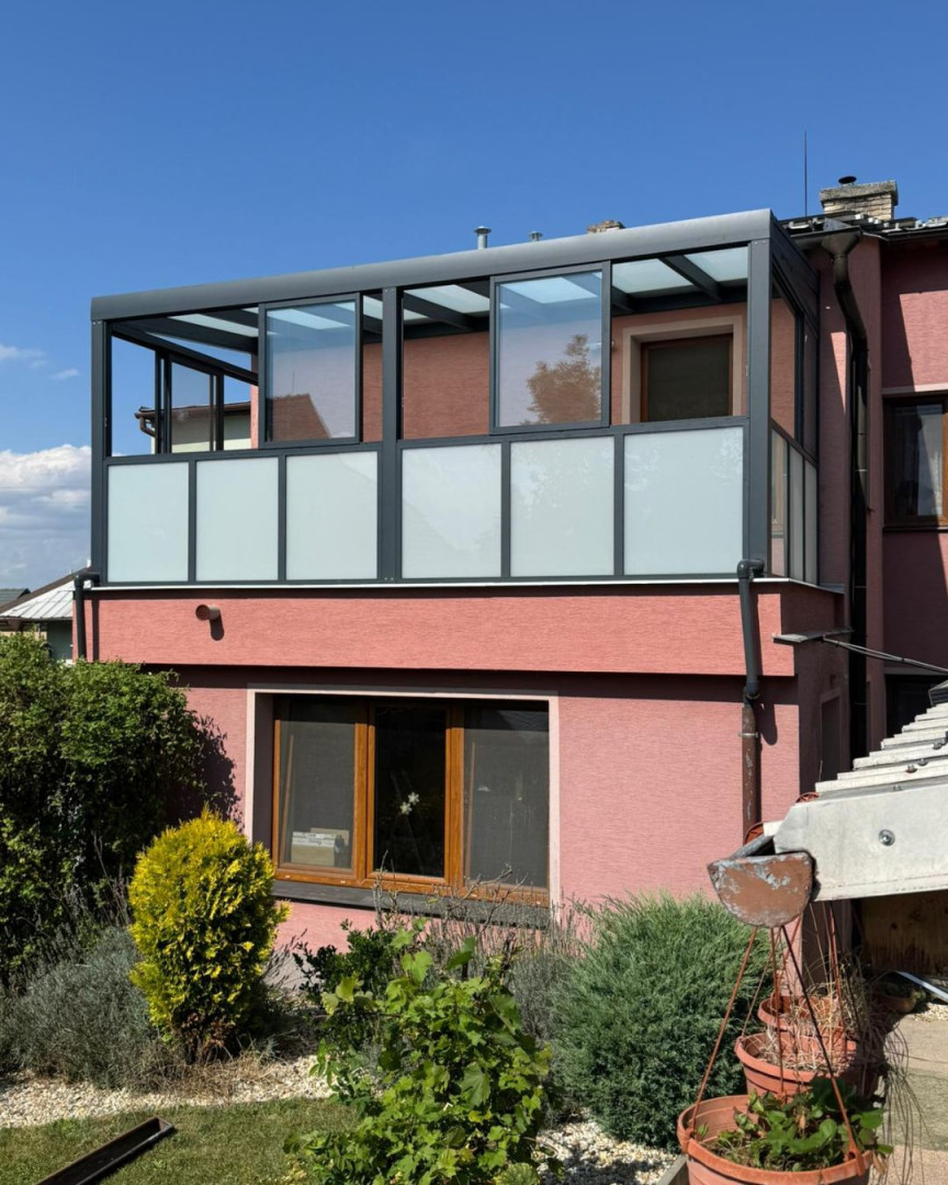 Moderní prosklená pergola s tmavě šedým hliníkovým rámem a matným sklem na balkóně rodinného domu.