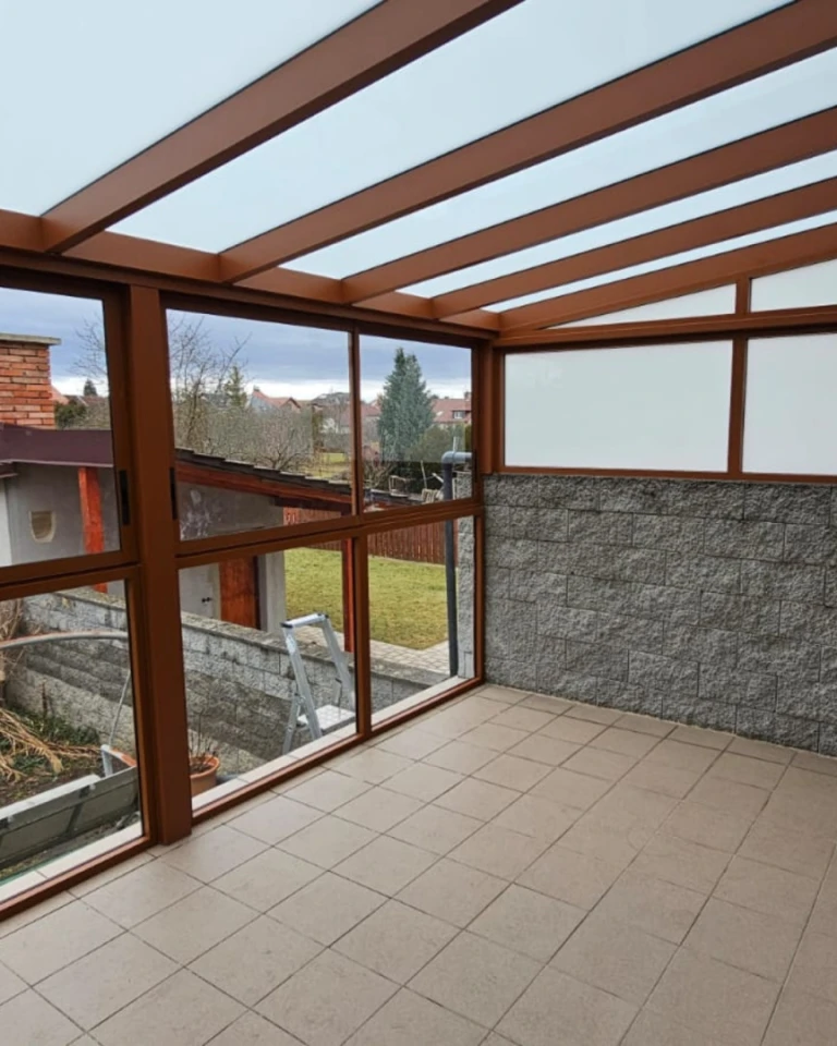 Prosklená hliníková pergola s hnědou konstrukcí a výhledem do zahrady
