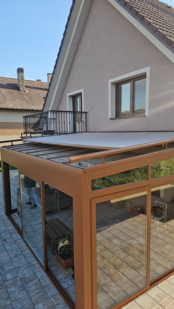 Prosklená zimní zahrada s hnědou konstrukcí a výsuvnou markýzou na terase rodinného domu