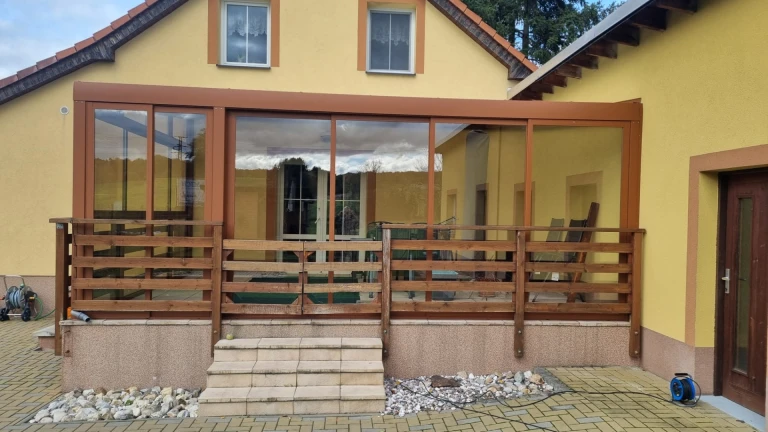 Moderní zimní zahrada s hnědou konstrukcí, prosklenými stěnami a dřevěným zábradlím před žlutým domem.